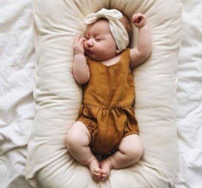 Baby nest bed in beige cotton with cushioned sides; newborn wearing a brown romper.