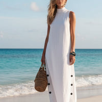 White sleeveless beach dress with side buttons, paired with woven bag on sandy shore