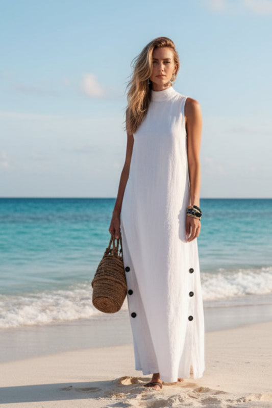 White sleeveless beach dress with side buttons, paired with woven bag on sandy shore