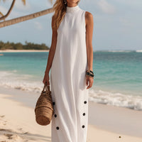 Woman in white sleeveless beach dress with black buttons, holding woven bag on sandy shore