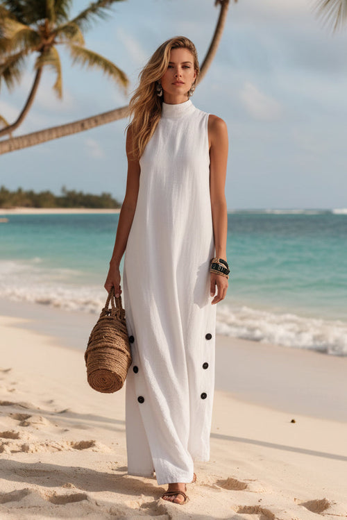 Woman in white sleeveless beach dress with black buttons, holding woven bag on sandy shore