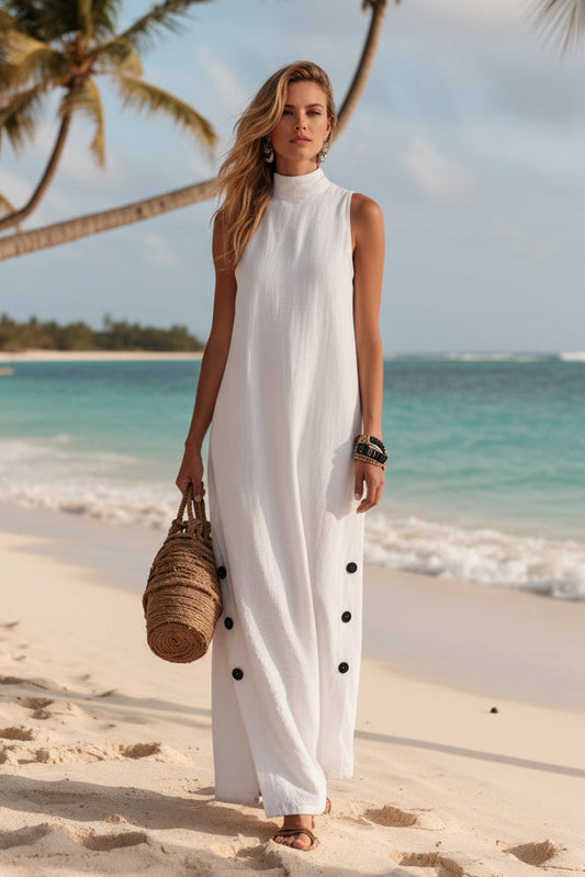 Woman in white sleeveless beach dress with black buttons, holding woven bag on sandy shore