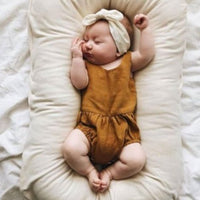 Baby nest bed in beige cotton with cushioned sides; newborn wearing a brown romper.